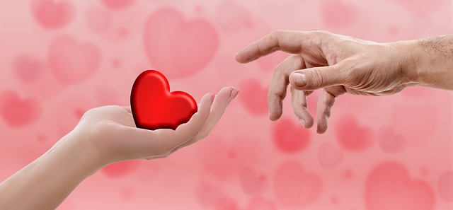 A red heart held in a hand, and being touched by someone's finger.