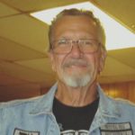 a grey haired man with glasses wearing a denim vest with many patches and a black T shirt with a circular standing in front of a blurred inside scene