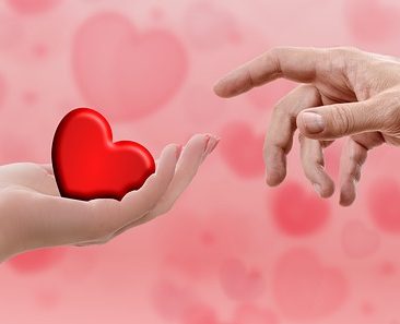 A red heart held in a hand, and being touched by someone's finger.