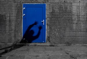 Grey brick wall and cement ground with a bright blue door in the middle of the wall. A shadow of a person reaches for the handle on the door.