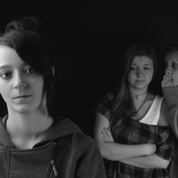 1 woman dressed in dark clothes with a sad face. in the background are two more women . One of them is whispering into the other's ear.