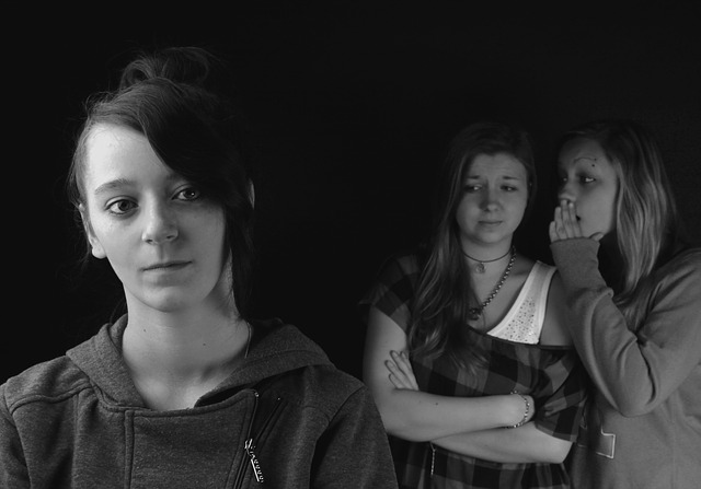 1 woman dressed in dark clothes with a sad face. in the background are two more women . One of them is whispering into the other's ear.